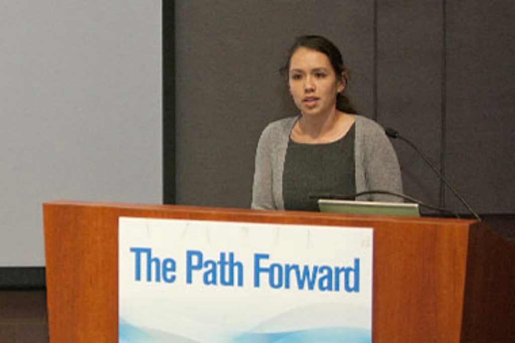 A woman speaks behind a podium with banner "The Path Forward"